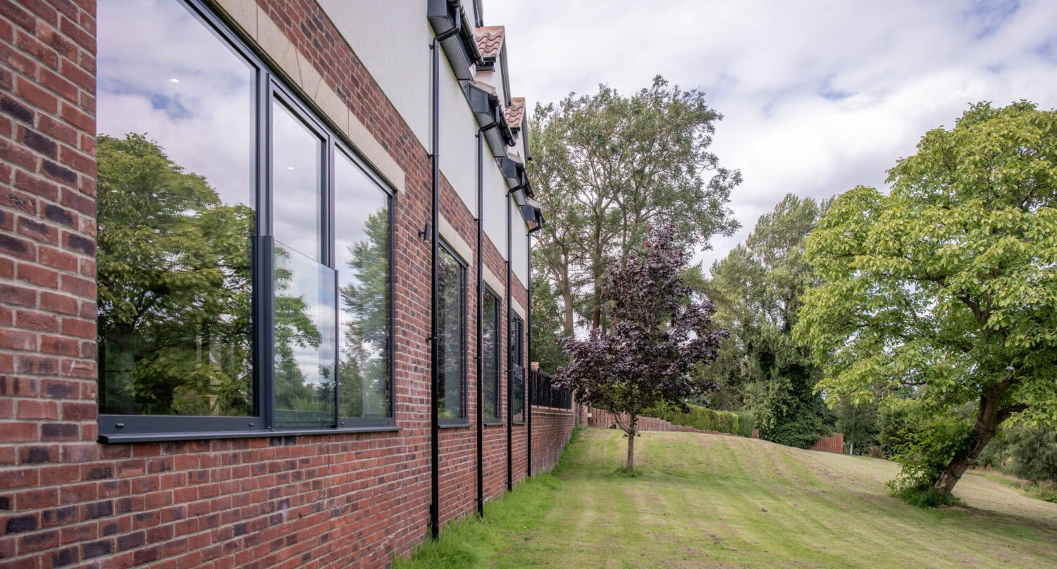A modern brick building with large windows and bespoke glass solutions reflects green trees outside. A well-kept lawn and several trees stretch alongside the building under a partly cloudy sky.