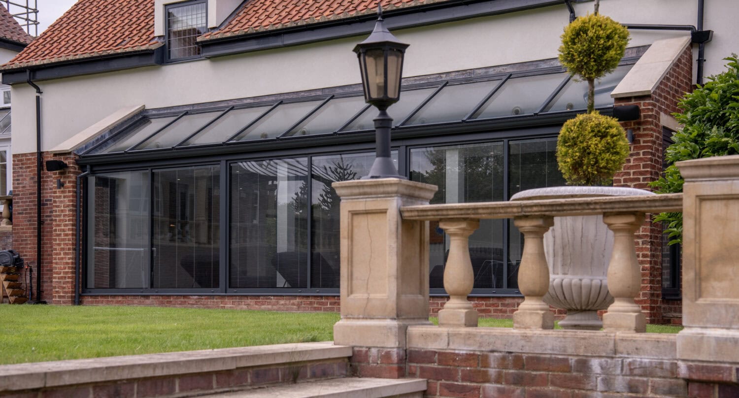 A modern glass conservatory extension with a sloped roof and sliding doors, attached to a brick house, viewed from a stone staircase with a balustrade and decorative topiary in the foreground.