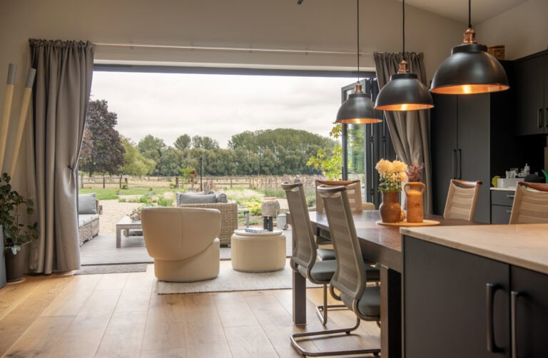 Modern open-plan kitchen and dining area with pendant lights, leading to a cozy patio with outdoor seating; large sliding doors reveal a scenic garden and trees beyond.