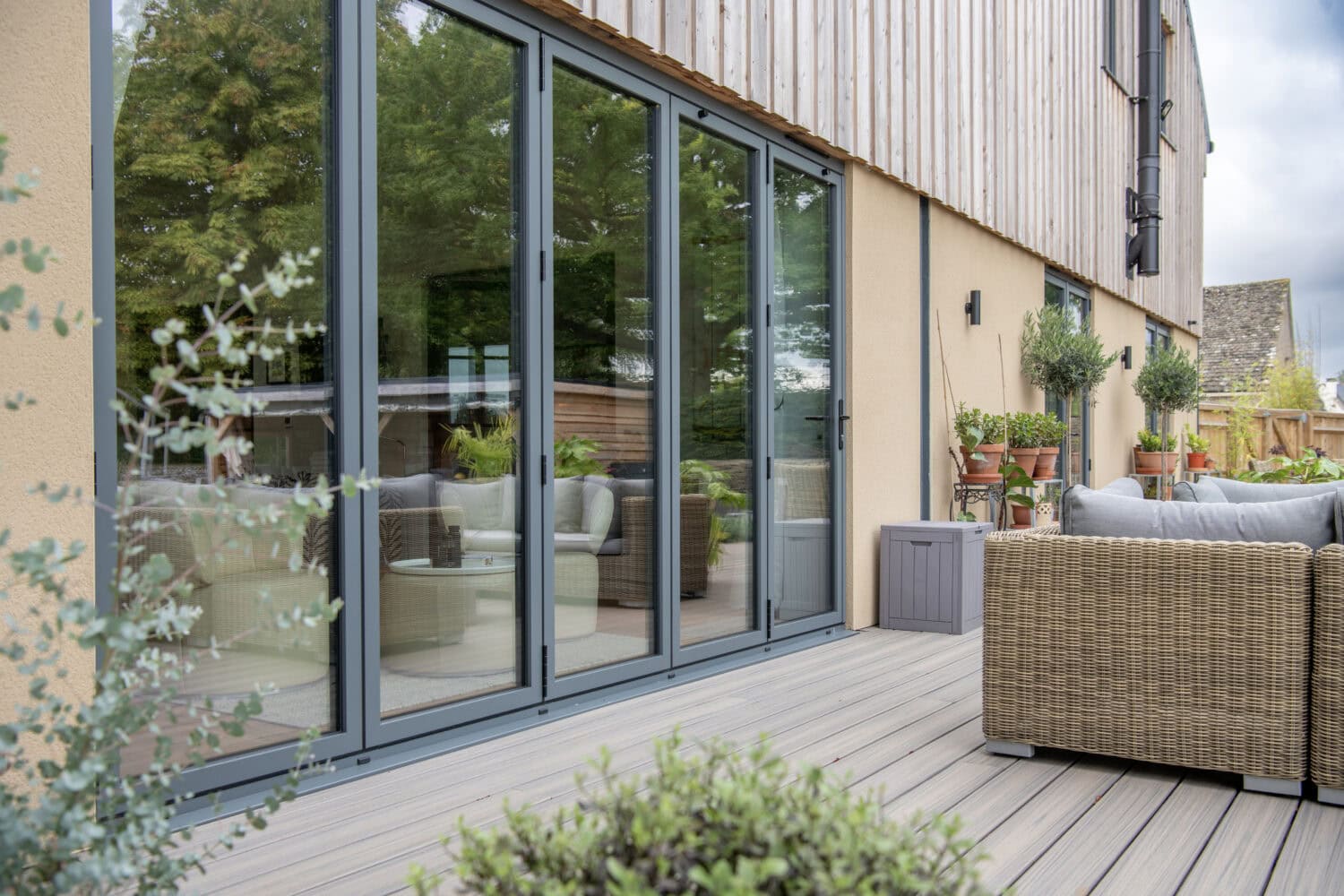 Modern patio with wicker furniture, potted plants, and decking, adjacent to a house with large glass sliding doors and optional glass roofing that reflects lush greenery from the garden.