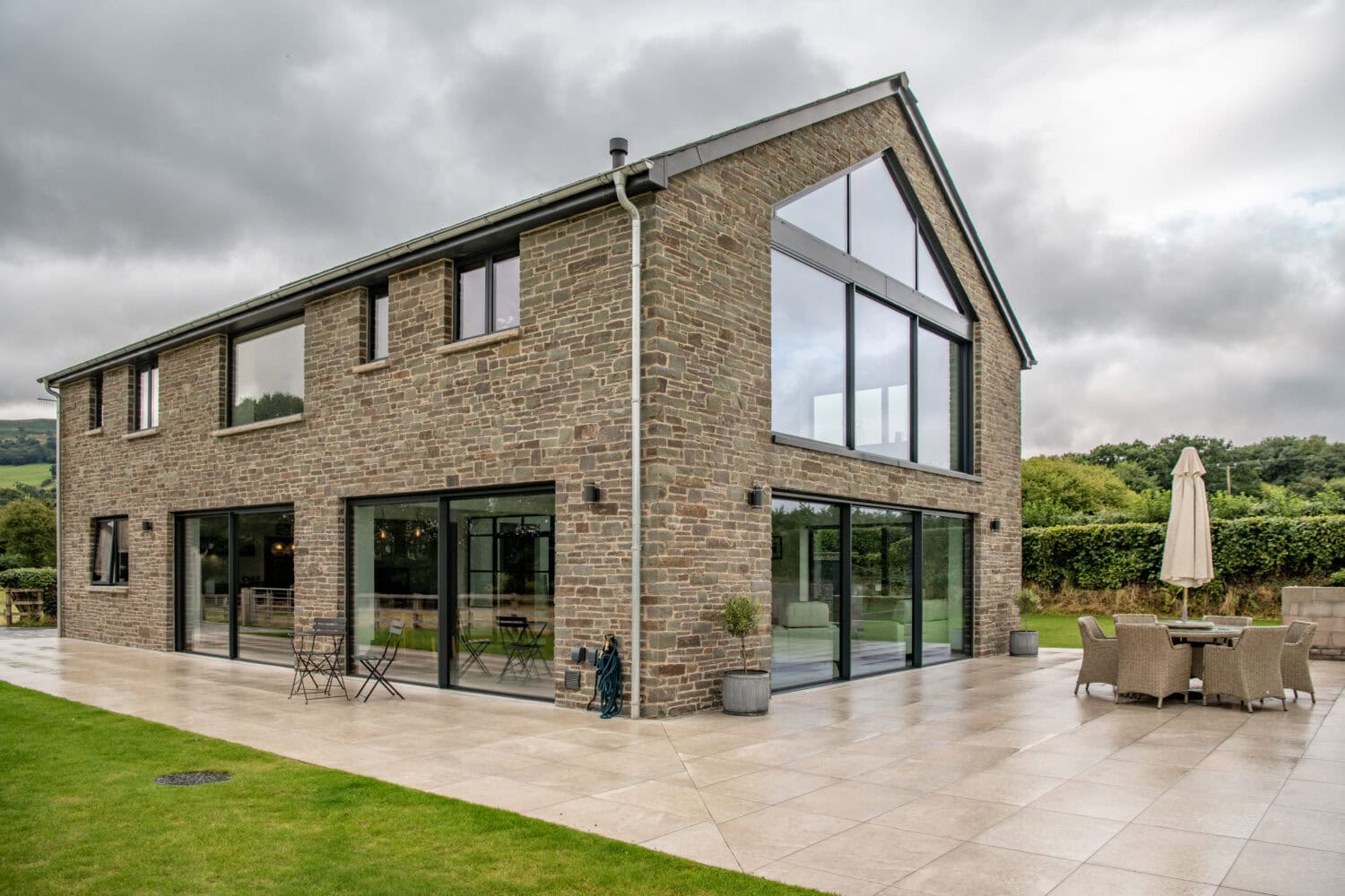 A modern two-story house with large glass windows, sliding doors, and stone exterior, set on a spacious patio with outdoor seating and surrounded by green lawn and countryside.