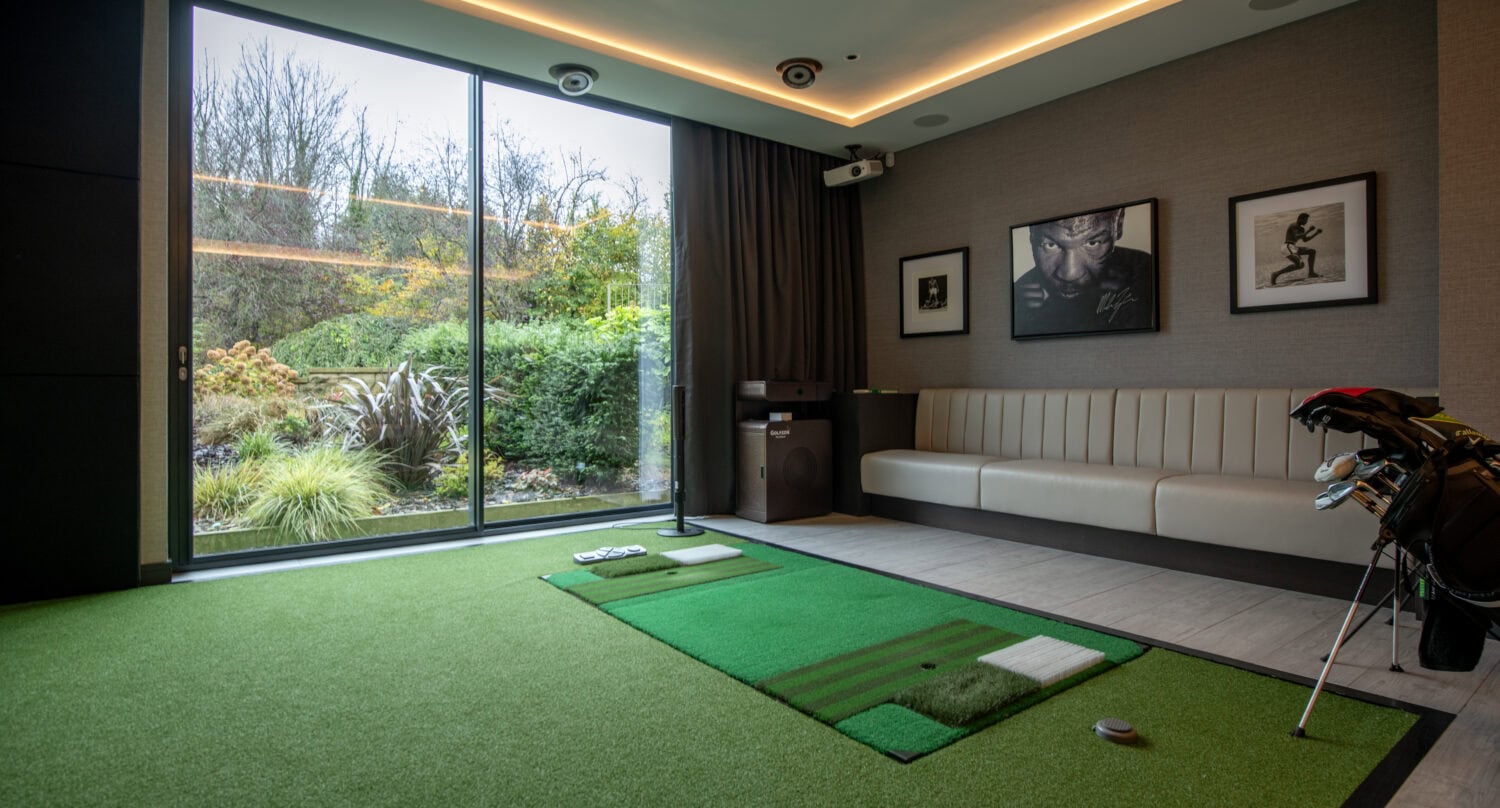 Modern indoor golf room with a green turf putting area, golf equipment, a white cushioned bench, framed sports photos on the wall, and large sliding glass doors showing a garden outside.