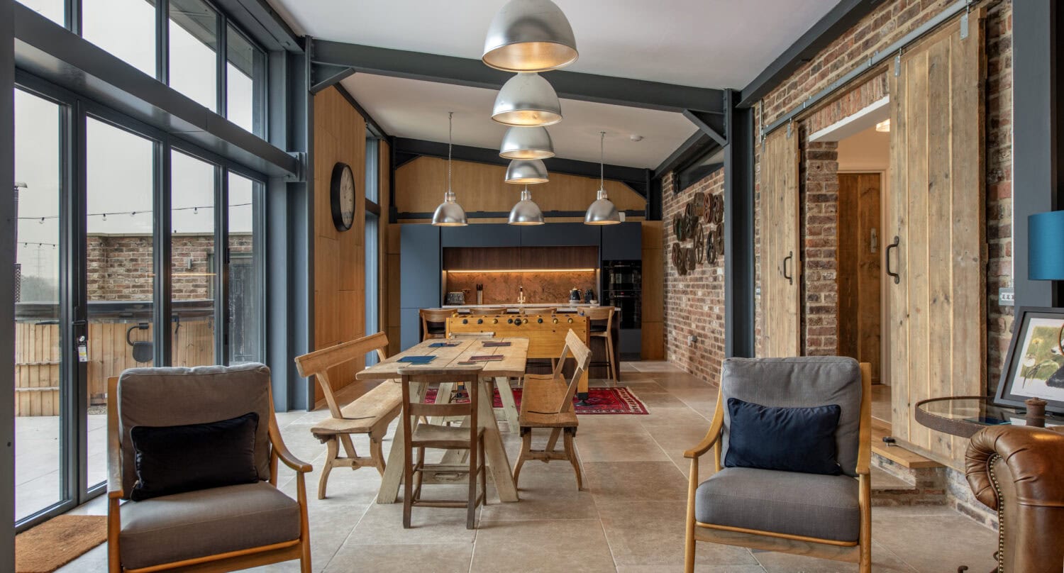Modern open-plan kitchen and dining area with large windows, industrial-style lighting, exposed brick walls, wooden furniture, a red rug, and bifold doors, creating a cozy yet contemporary atmosphere.