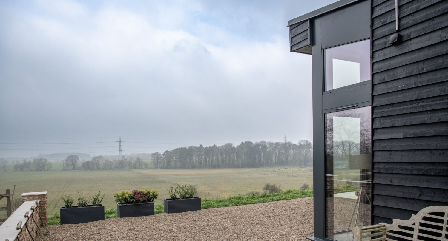 Modern black wooden building with bifold doors and large glass windows next to a gravel patio, benches, and planters, overlooking a grassy field and distant trees under a cloudy sky.