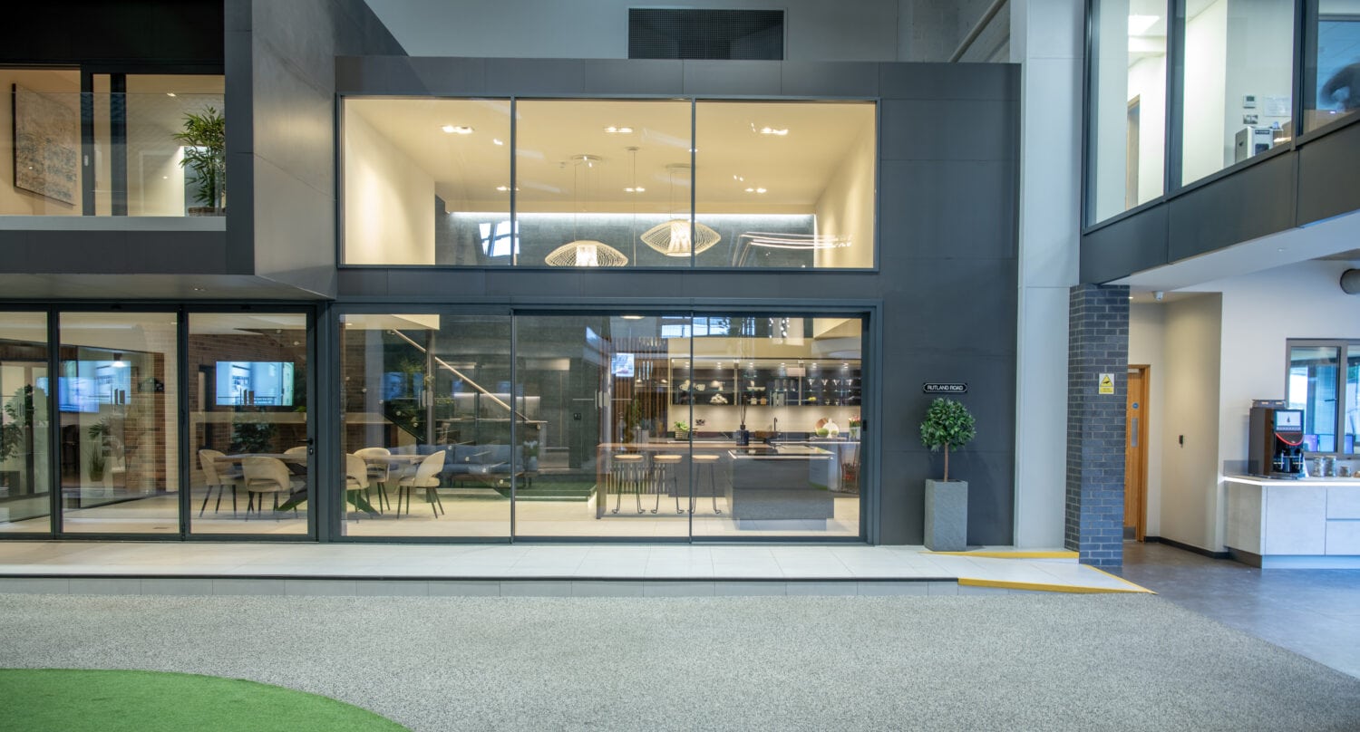 Modern office interior with large glass windows, open meeting spaces, and contemporary furnishings. Bespoke glass solutions include glass roofing upstairs with pendant lights, while downstairs offers seating areas and a visible reception or common area.