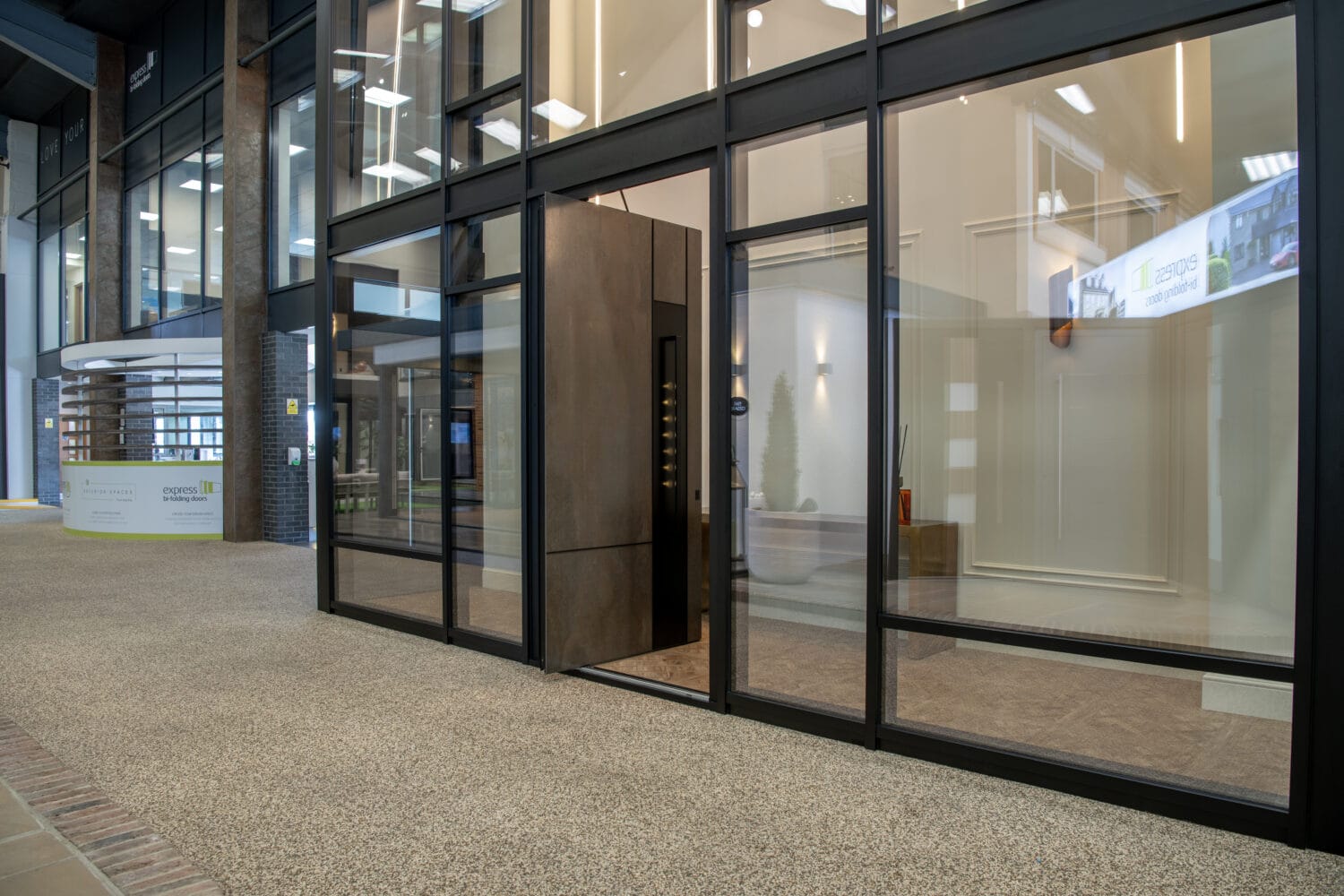 Modern glass office entrance with a brown front door, transparent walls, and carpeted floor in a building lobby. Reflections of ceiling lights are visible on the glass. A reception desk is seen in the background.