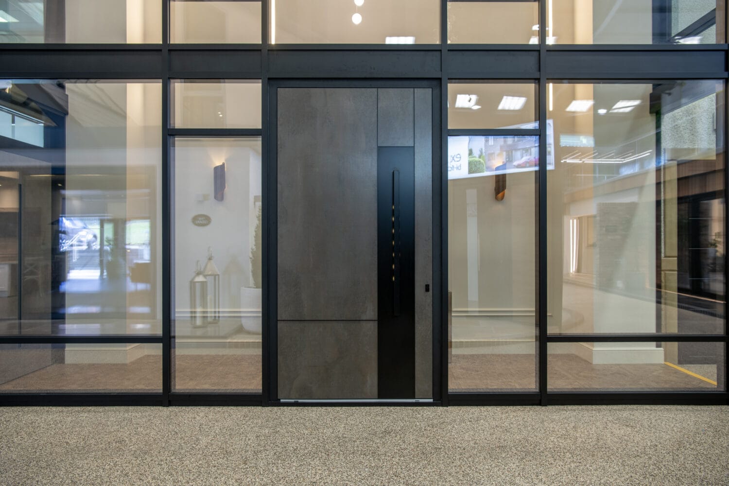 A modern building entrance with sliding doors and tall glass panels, featuring a large dark door. The bright interior showcases reflections of lights, indoor decor, and bespoke glass solutions throughout.