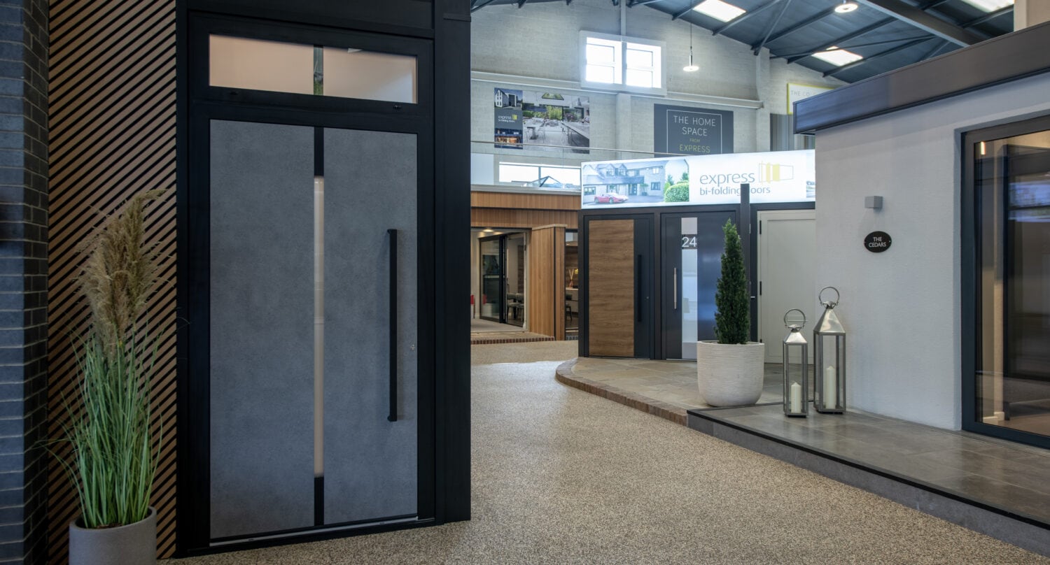 A modern indoor showroom displays various contemporary front doors and windows, with plants and decorative lanterns near the entrances under a high, industrial-style ceiling featuring sleek glass roofing.