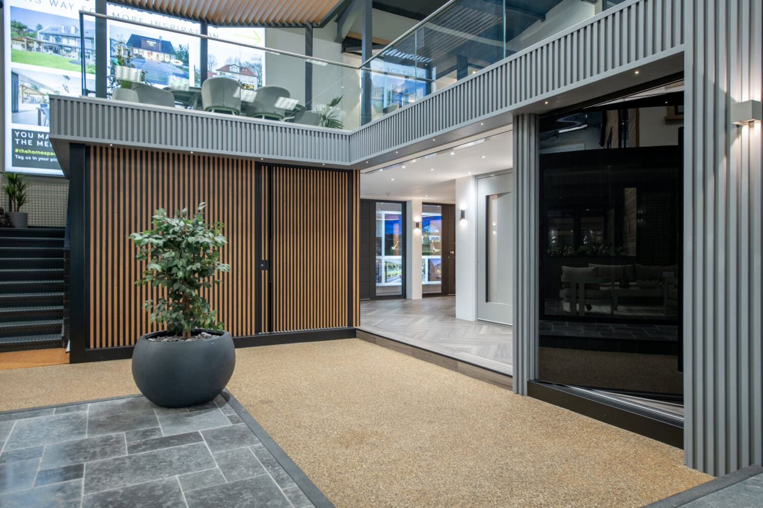 Modern indoor showroom with large windows, wooden paneling, a potted plant, tiled floor, sliding doors, and glass balcony railing. Light from digital displays and recessed lighting creates a bright, contemporary atmosphere.