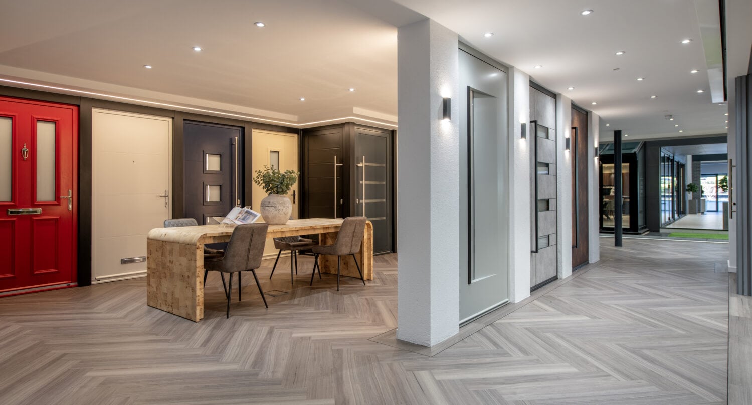 A modern showroom displays various front doors and sliding doors, with a table and chairs arranged in the center on a herringbone-patterned floor, under bright ceiling lights.