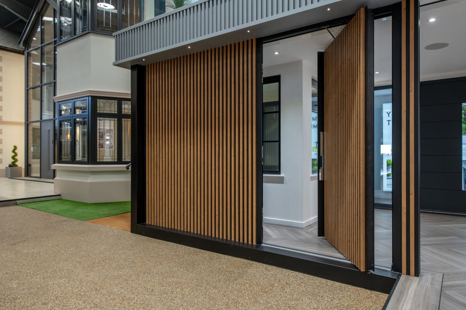 Modern building entrance with vertical wooden slats forming a pivoting door, partially open, revealing a stylish interior with large windows and sliding doors, plus a mix of textured flooring and greenery outside.