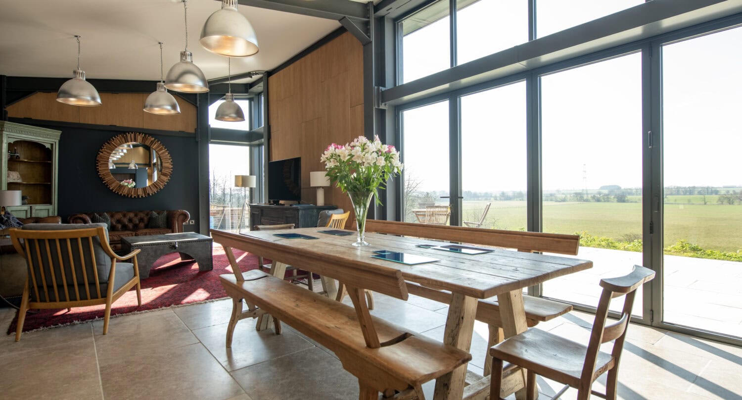 A modern, open-plan dining and living area with large floor-to-ceiling windows, sliding doors, wooden tables and benches, pendant lights, a vase of flowers, and views of green fields outside.