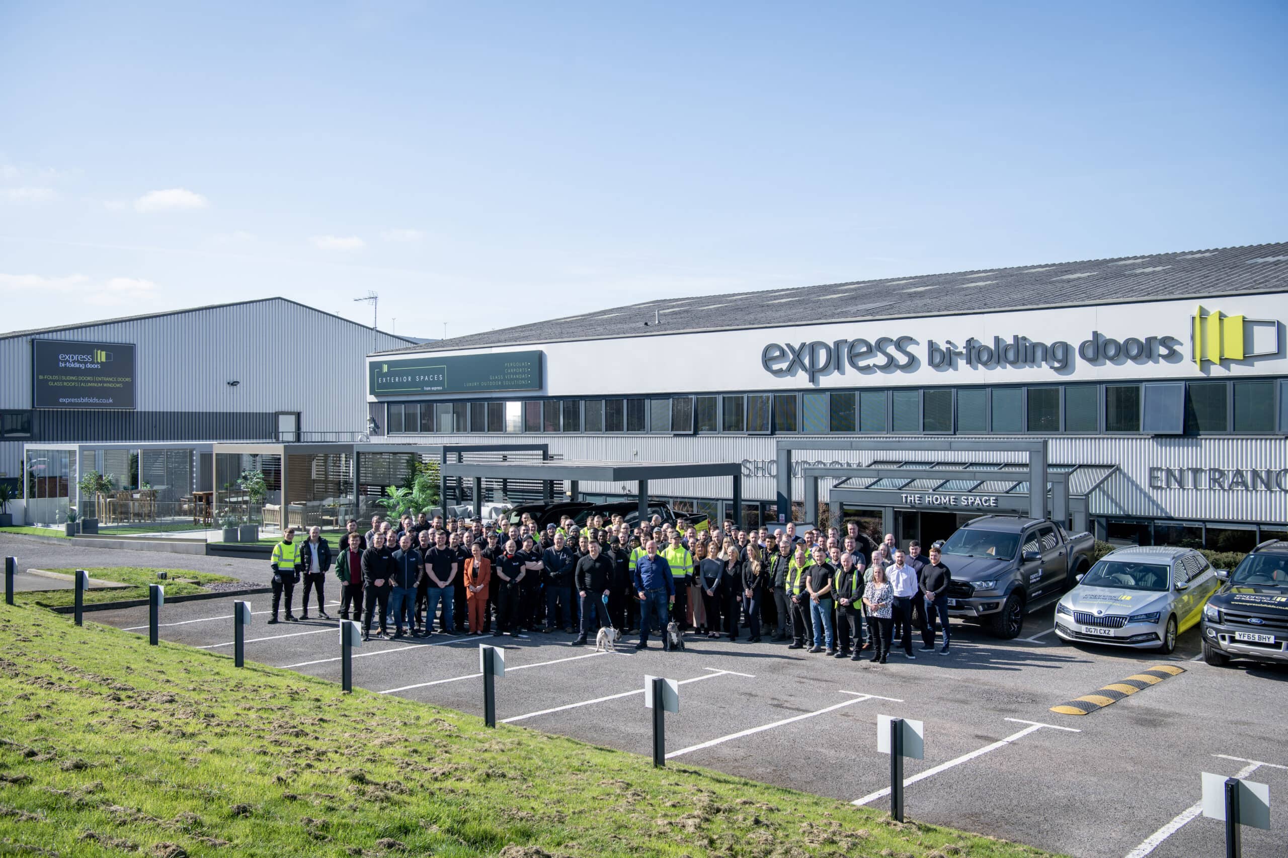 A large group of people stands together in front of the Express Bi-Folding Doors building and showroom, known for stunning bifold doors, with several cars parked nearby and clear blue skies overhead.
