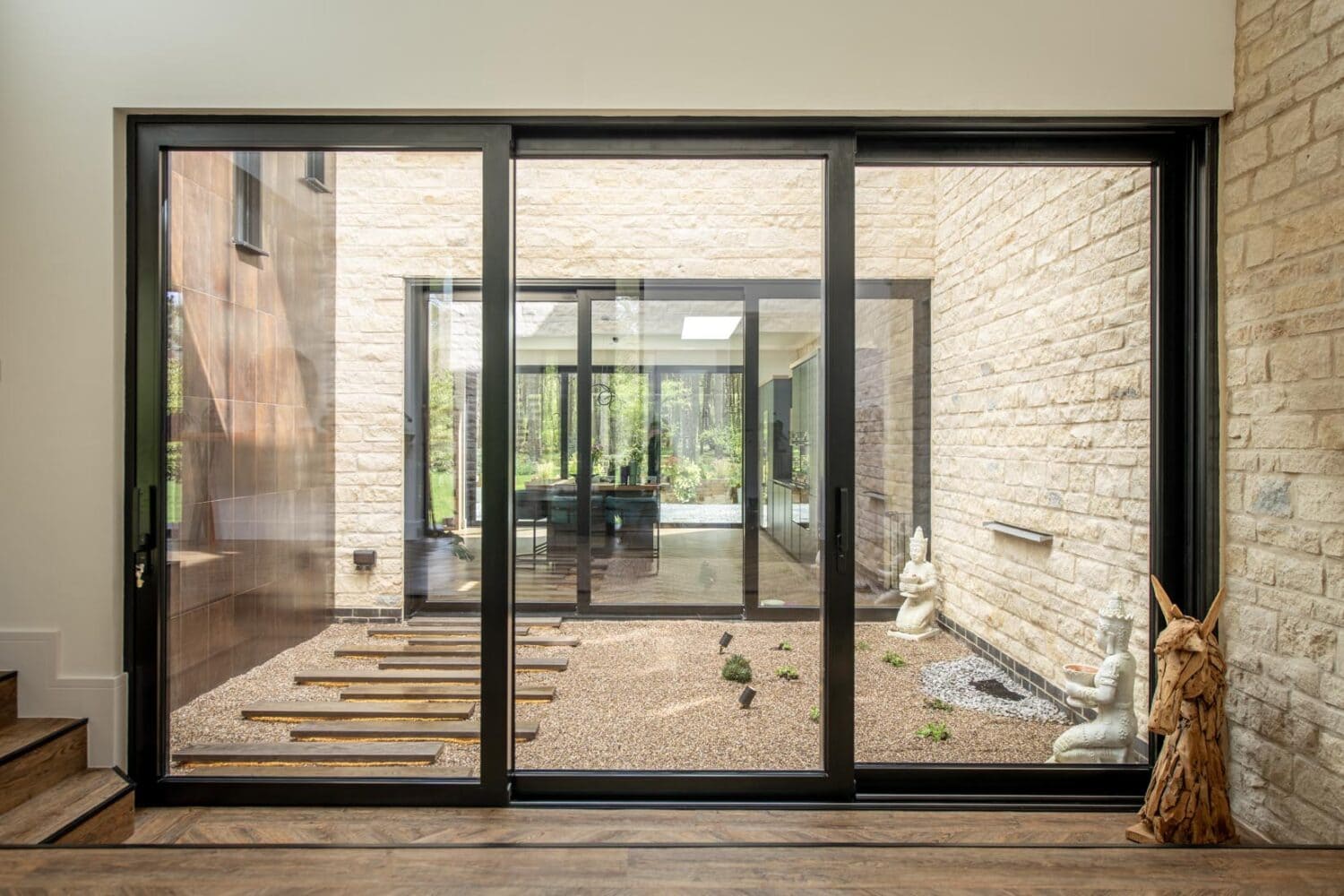 A modern indoor courtyard with gravel, stepping stones, small plants, and white stone statues, viewed through bespoke glass solutions like large bifold doors from inside a house with light-colored brick walls.