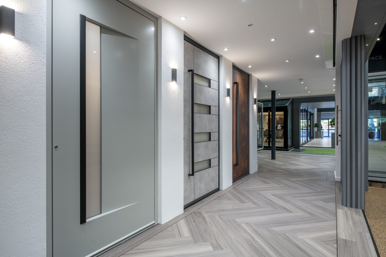 A modern hallway with gray herringbone-patterned flooring, contemporary doors on the left, wall lights, and bespoke glass solutions revealing a green outdoor area at the end. The space is clean and brightly lit.