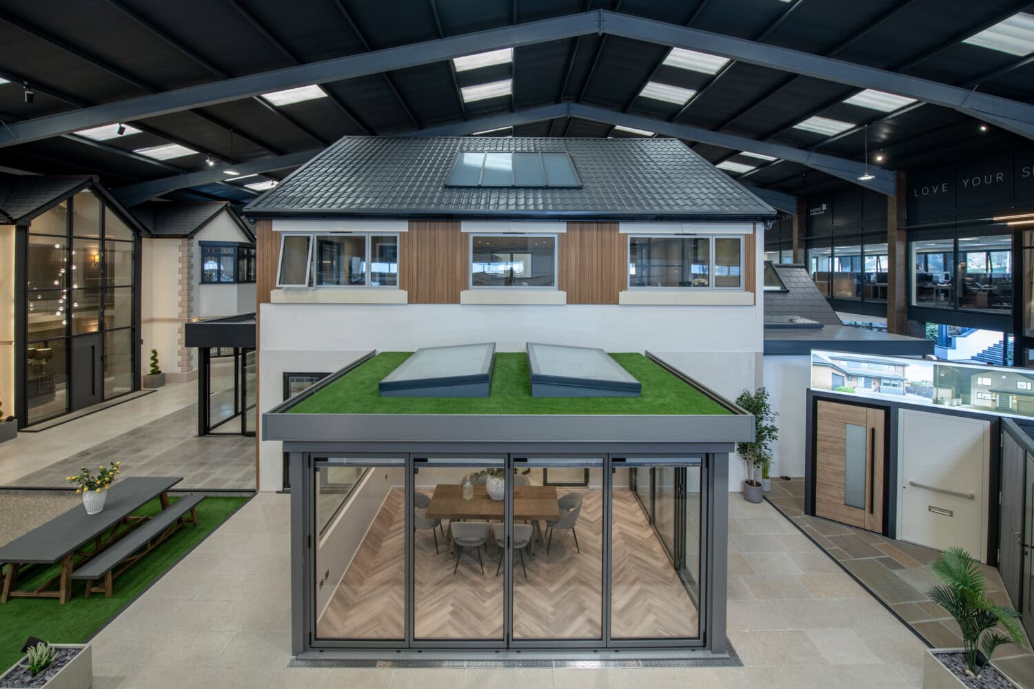 Modern showroom interior displaying a two-story house model with a glass-walled extension featuring bifold doors to the dining area, artificial grass roof, and skylights; other model homes visible in the background.
