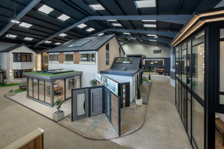 A spacious indoor showroom displays several modern house models with large windows, bespoke glass solutions for front doors, and outdoor patio areas, all under a high ceiling with skylights.