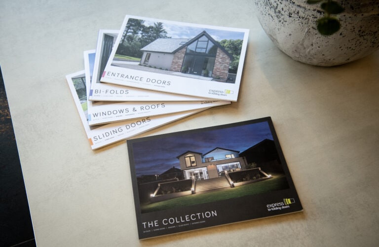 A stack of brochures about windows and doors lies on a table, with the top one titled The Collection showing a modern house at night. Other brochures mention front doors, bi-fold doors, windows & roofs, and sliding doors.