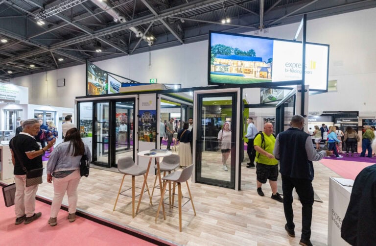 A trade show booth displays large glass front doors and windows; several people are walking around, talking, and examining the products in a spacious exhibition hall with high ceilings and bright lighting.