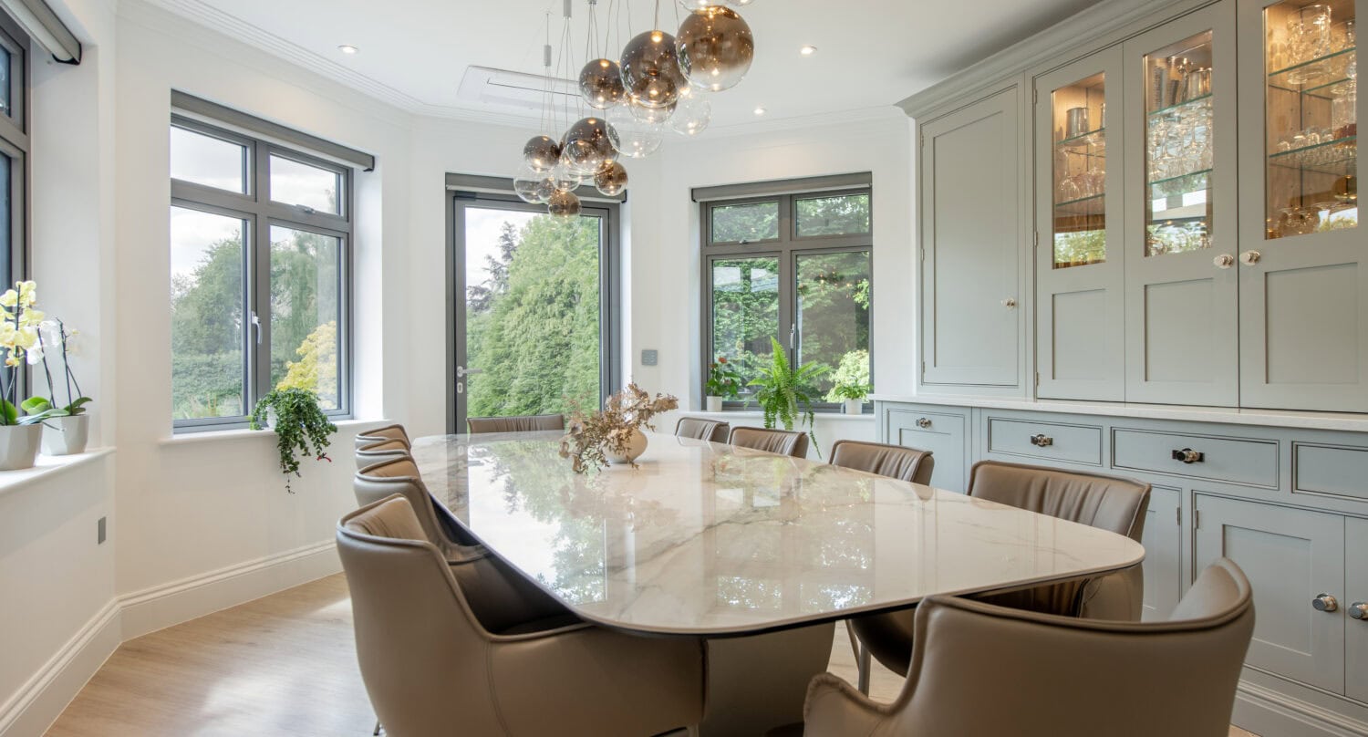 A modern dining room with a marble table, eight tan leather chairs, pendant lights, large windows with garden views, and built-in cabinets with glass doors displaying glassware.