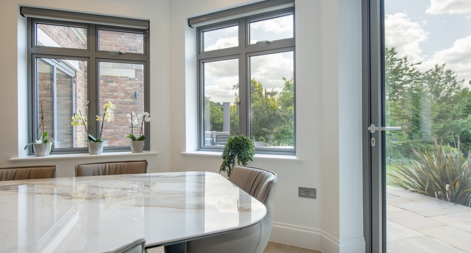 Modern dining area with a marble table, beige chairs, large windows, and glass door overlooking a green garden. Several potted plants sit on the windowsills, and natural light fills the bright, clean room.