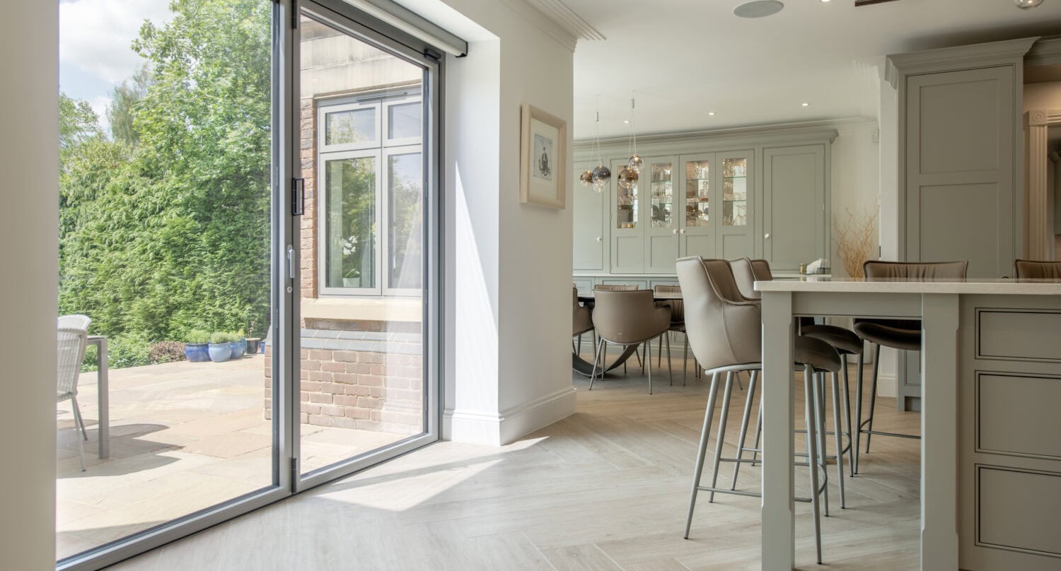 Modern kitchen and dining area with light wood floors, neutral cabinetry, and a large glass sliding door opening to a sunny patio with outdoor seating and lush greenery.