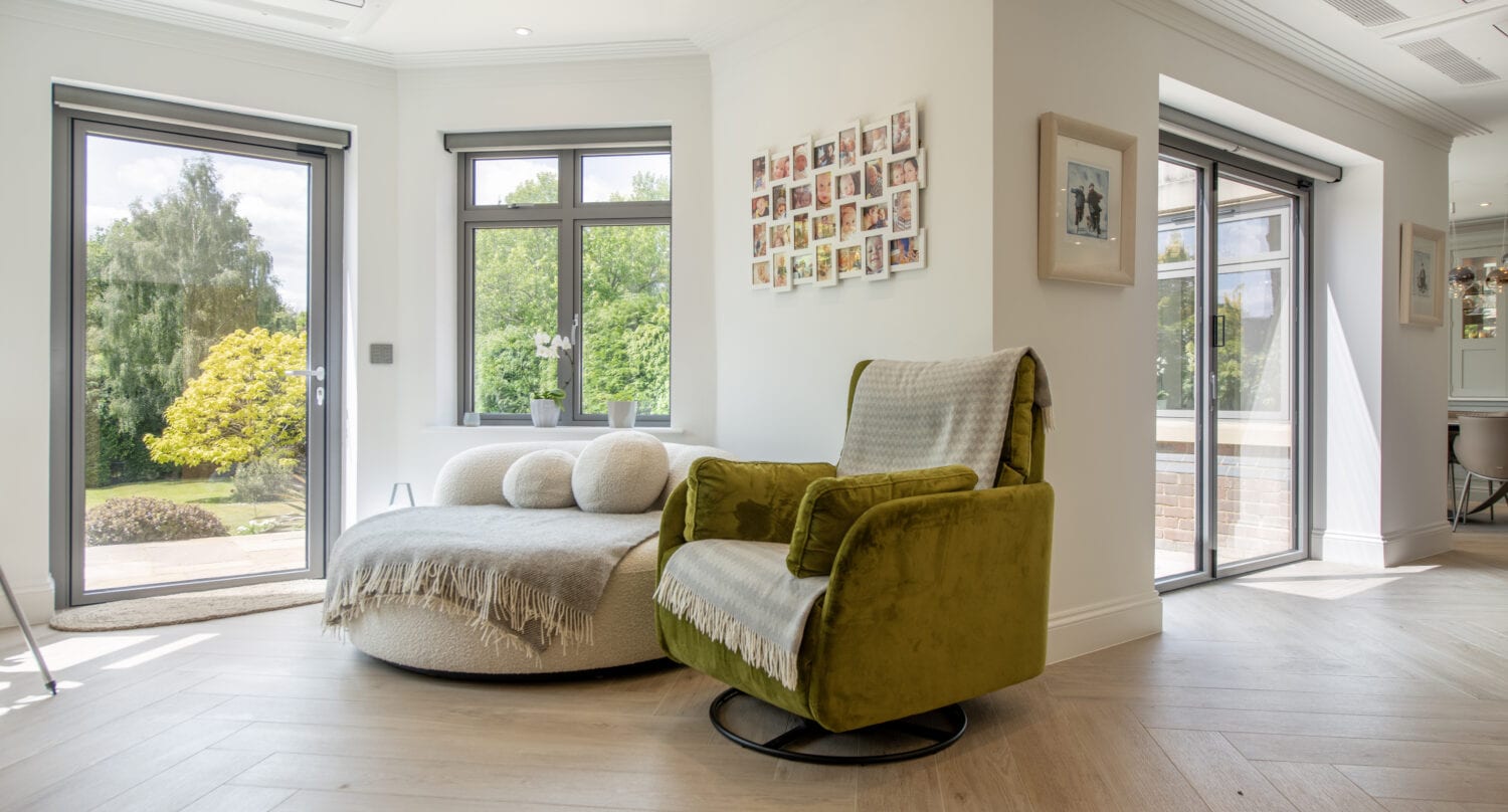 A cozy, modern living space with large windows, a green armchair, a round beige chaise lounge with cushions and a throw, light wood flooring, and a photo collage on the wall, overlooking a lush green garden.