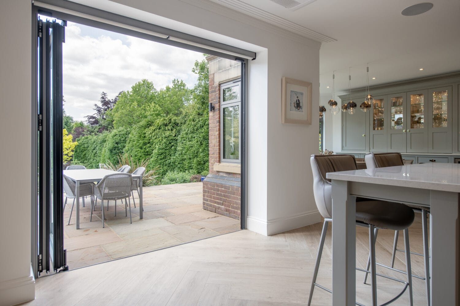 Modern kitchen with an island and bar stools opens to a patio through large folding glass doors. Outdoor dining table and chairs are set on a paved terrace, overlooking a lush green garden.
