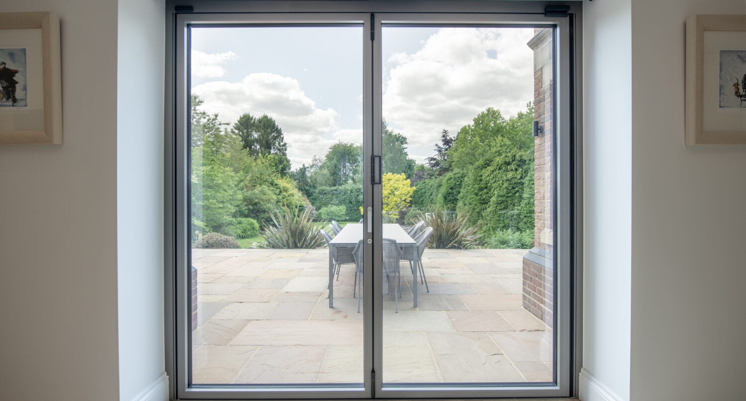 A large glass sliding door opens to a stone patio with a table and chairs, overlooking a lush garden with green trees and bushes under a partly cloudy sky.
