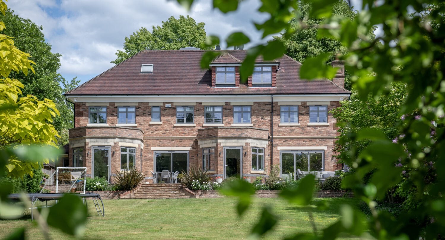 A large, two-story brick house with multiple windows and a well-kept lawn is framed by leafy green branches, under a partly cloudy sky.
