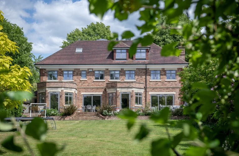 A large, two-story brick house with multiple windows and a well-kept lawn is framed by leafy green branches, under a partly cloudy sky.