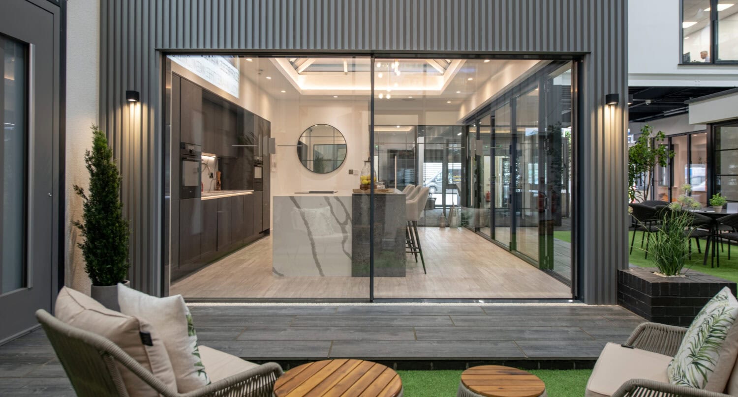 Modern indoor kitchen and dining area viewed through bespoke glass solutions with large sliding doors, opening onto a cozy outdoor seating area with chairs, a small table, and lush greenery in the foreground.