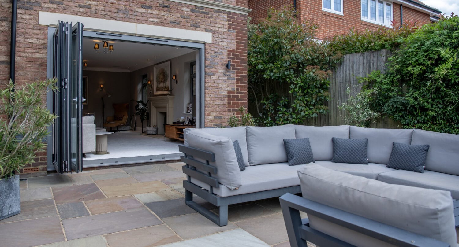 A modern outdoor patio with gray cushioned seating is shown next to a brick house with large folding doors open to a stylish living room inside. The patio has stone tiles and is surrounded by greenery and a wooden fence.