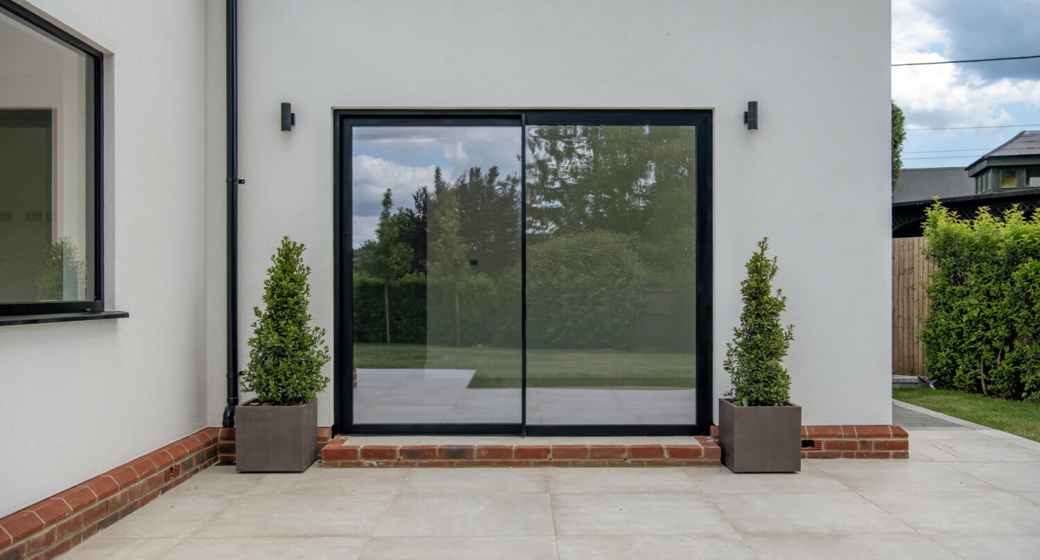 Modern black-framed sliding bi-fold doors installed on a white house exterior, opening to a garden with lush greenery, enhancing natural light and indoor-outdoor flow.