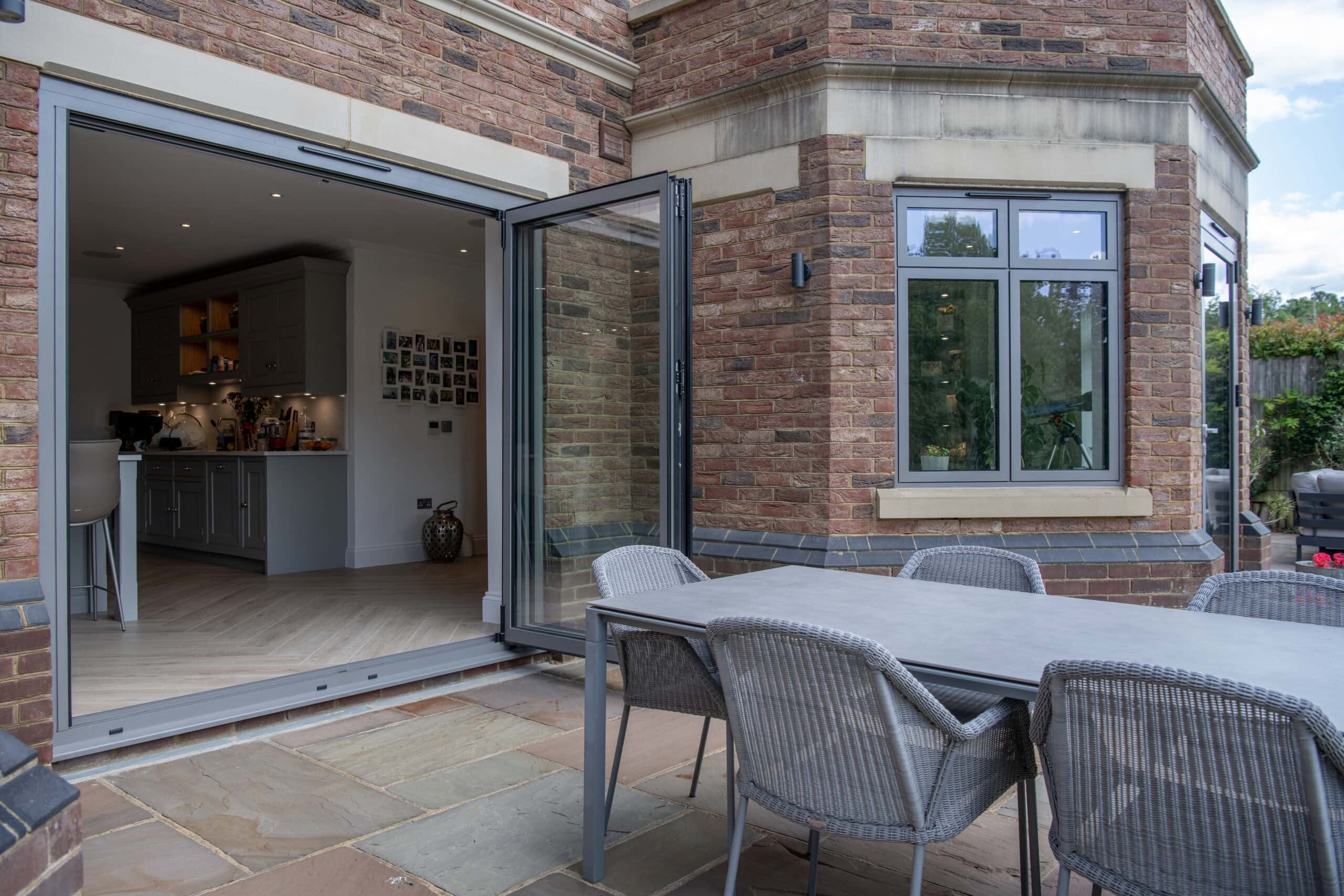 A modern brick house with open folding glass doors leading from the kitchen to a patio. On the patio is a gray dining table with six woven chairs. The kitchen interior and some outdoor greenery are visible.