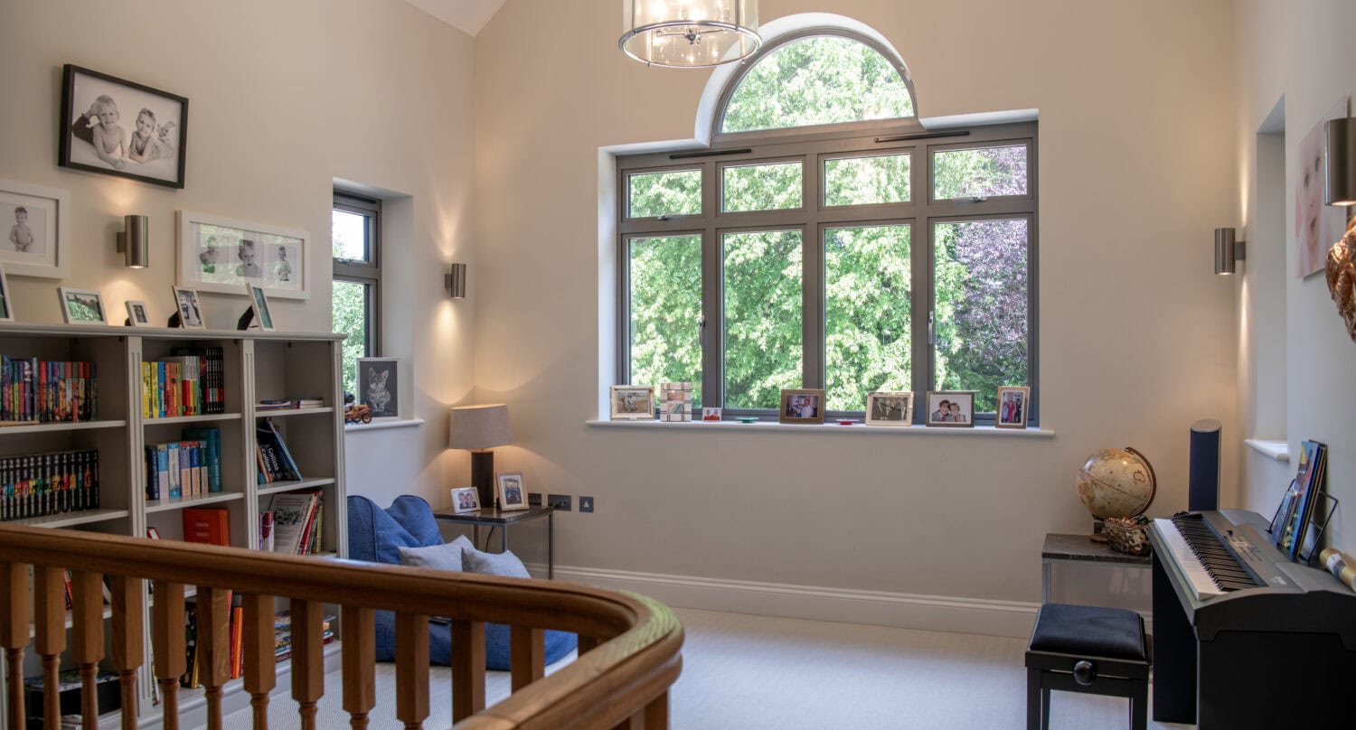A bright room with large arched windows, a piano, bookshelves filled with books and photos, a blue chair, a globe, and framed pictures on display. Natural light illuminates the beige walls and carpet.