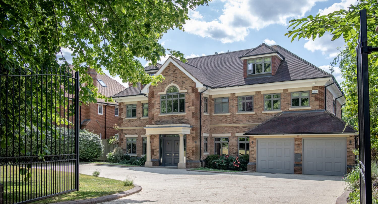 A large, modern brick house with a symmetrical facade, tall windows, and double garage, surrounded by greenery and partially shaded by trees, viewed from behind an open black metal gate on a sunny day.