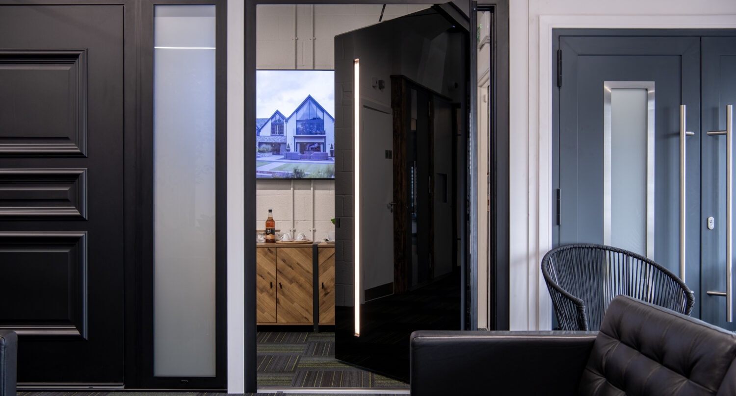 A modern office lobby with black leather seating, a dark door slightly open revealing a TV displaying house images, sliding doors leading to another area, a wood cabinet, and a black chair in the corner.