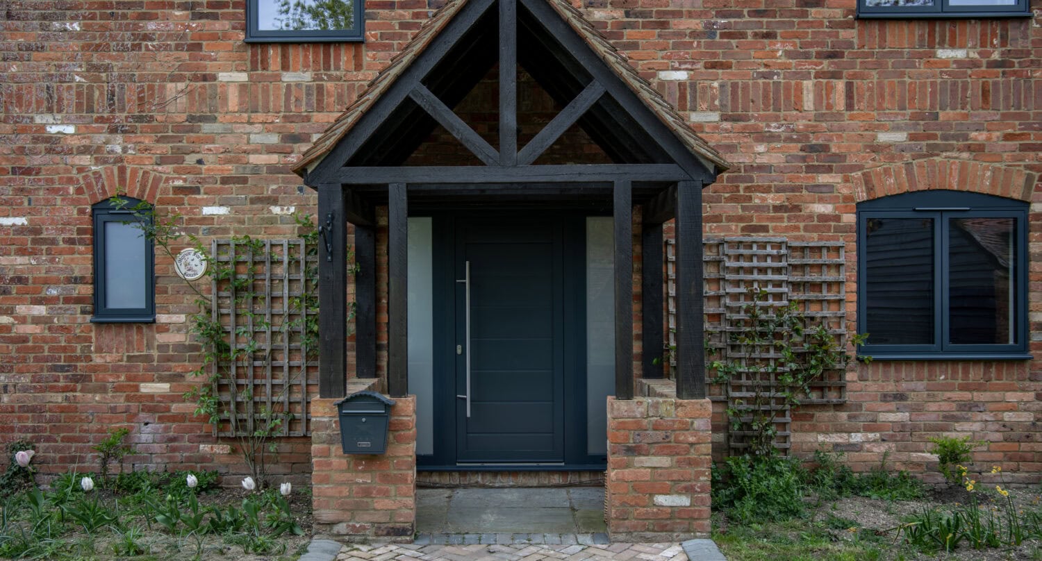Stylish black front door with a wooden canopy, set in a brick house with garden plants and windows, ideal for enhancing home entrance appeal.