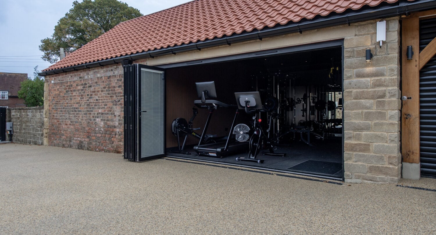 Modern garage with black bi-folding doors open to reveal gym equipment inside, set against a brick wall and red tiled roof, under a clear sky.