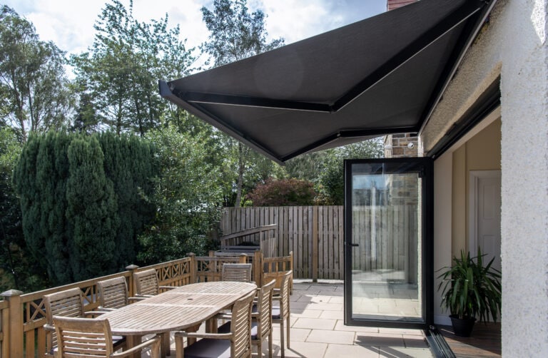 Modern outdoor patio featuring black bi-folding doors, a large canopy, and wooden dining furniture, ideal for outdoor living and solar shading solutions.