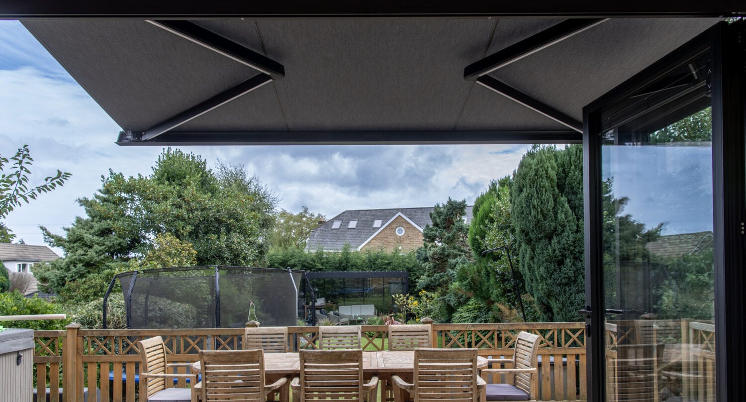 Modern outdoor patio with bi-folding doors, wooden dining table, and lush garden view, perfect for outdoor living and solar shading solutions.
