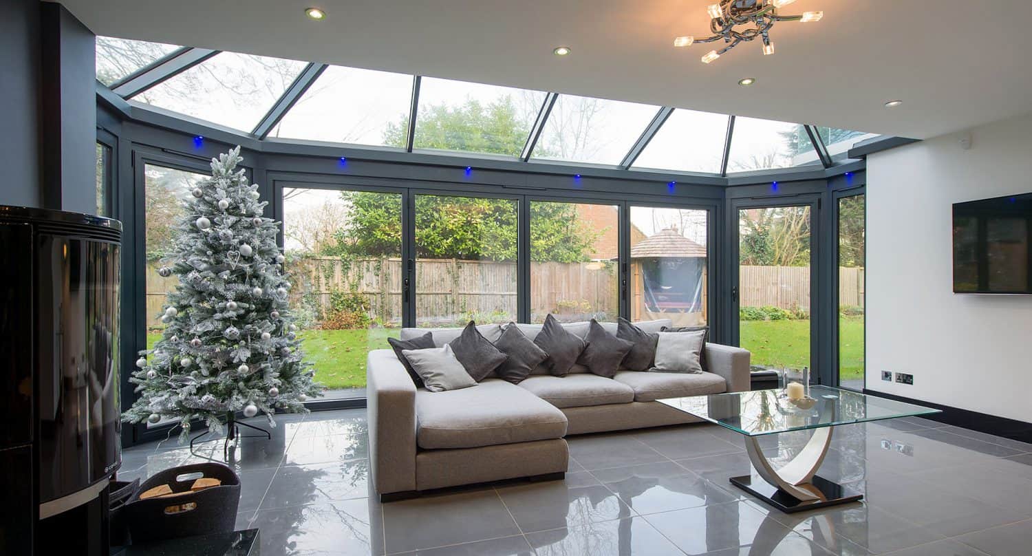 Modern conservatory with a grey sofa, glass coffee table, and frosted Christmas tree, featuring large windows and glass ceiling overlooking a green garden.