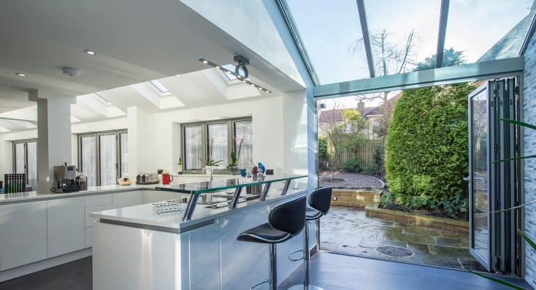 Modern kitchen with white cabinets and an island with two black bar stools, featuring bespoke glass solutions like large glass doors that open to a patio and lush green garden. Natural light fills the space through skylights and windows.