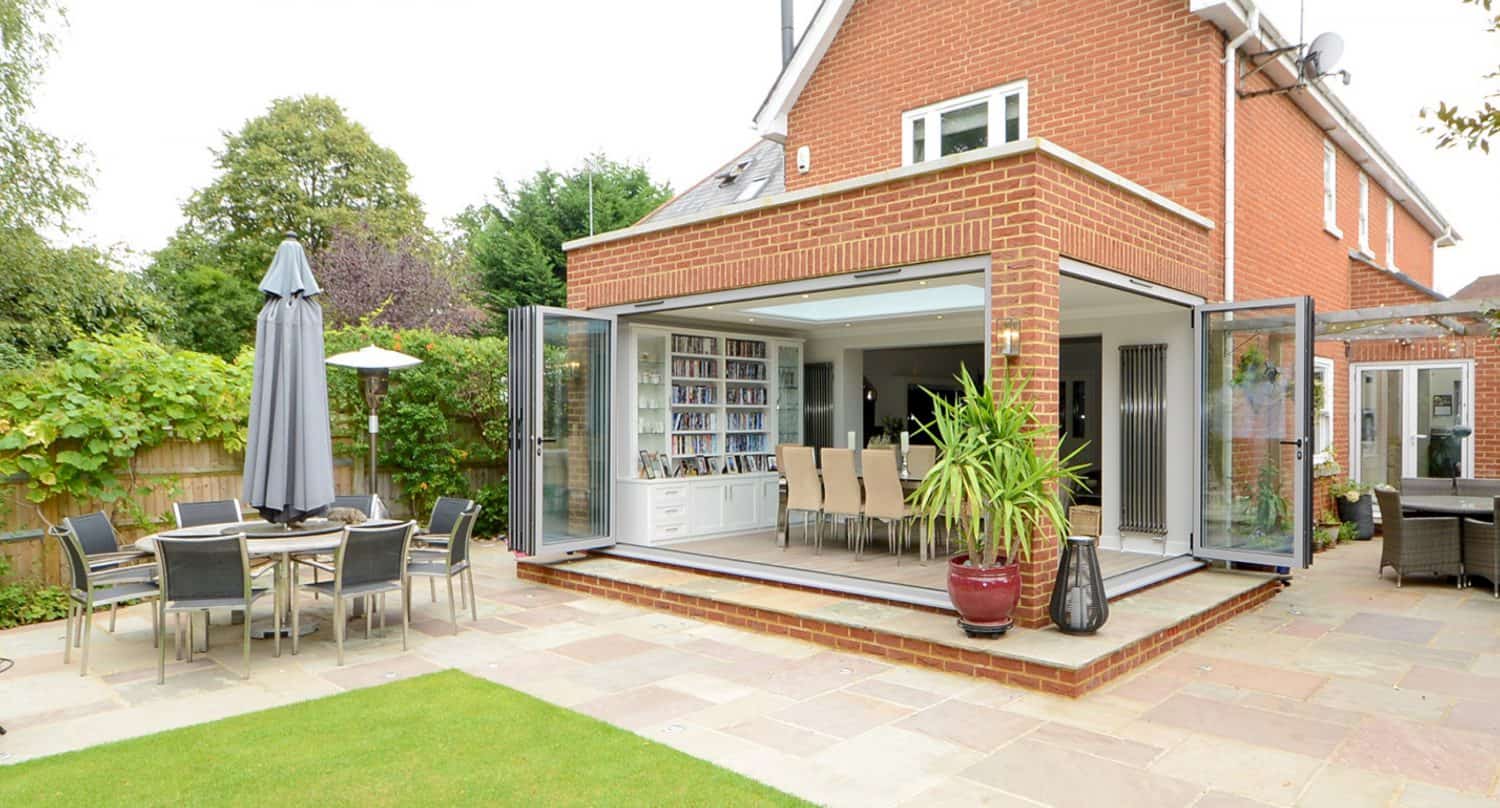 A modern brick house with large glass doors open to a patio. There’s a dining area inside, a table with chairs and umbrella outside, potted plants, and a well-kept lawn surrounded by greenery.