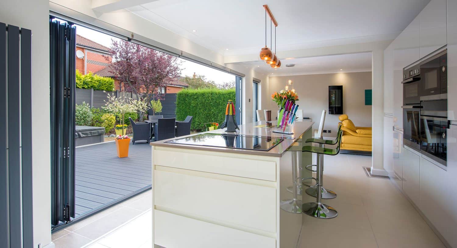 Modern kitchen with white island, built-in appliances, and pendant lights, opening onto a patio with outdoor seating and greenery through large folding glass doors.
