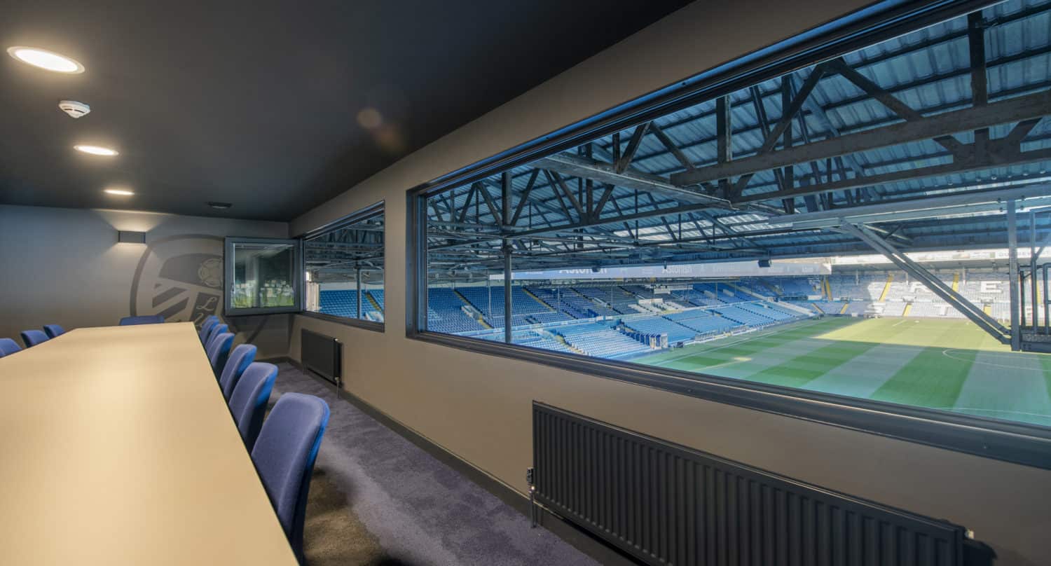 View from a private suite inside a stadium, featuring bespoke glass solutions with large windows and glass roofing, revealing blue chairs and a long table overlooking the empty blue seats and a well-lit green football pitch outside.