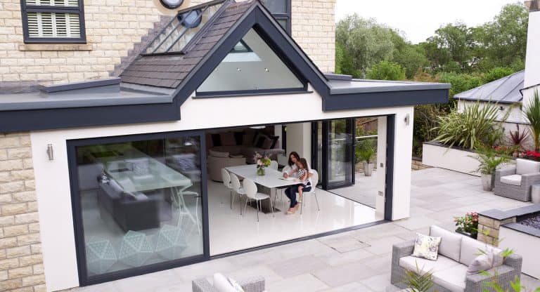 A modern house with large glass sliding doors opens to a patio. Inside, a woman sits at a dining table with a child, reading together. The kitchen and living area are visible; outdoor seating is arranged on the patio.