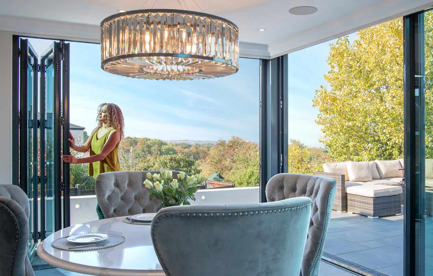 A woman opens bespoke glass bifold doors in a modern dining room with a round table and chandelier, leading to a spacious outdoor patio with seating and views of trees.