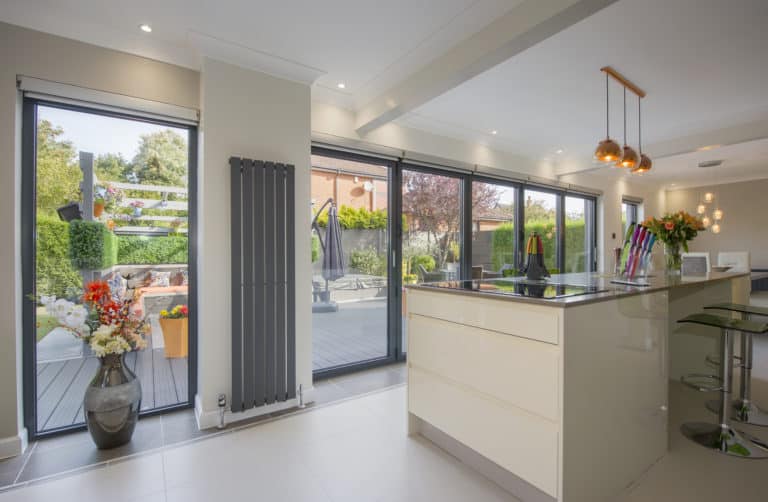 Modern kitchen with glossy island, bar stools, and pendant lights, opening onto a patio through large sliding doors; outdoor greenery and flowers can be seen, with decorative vases on display inside.